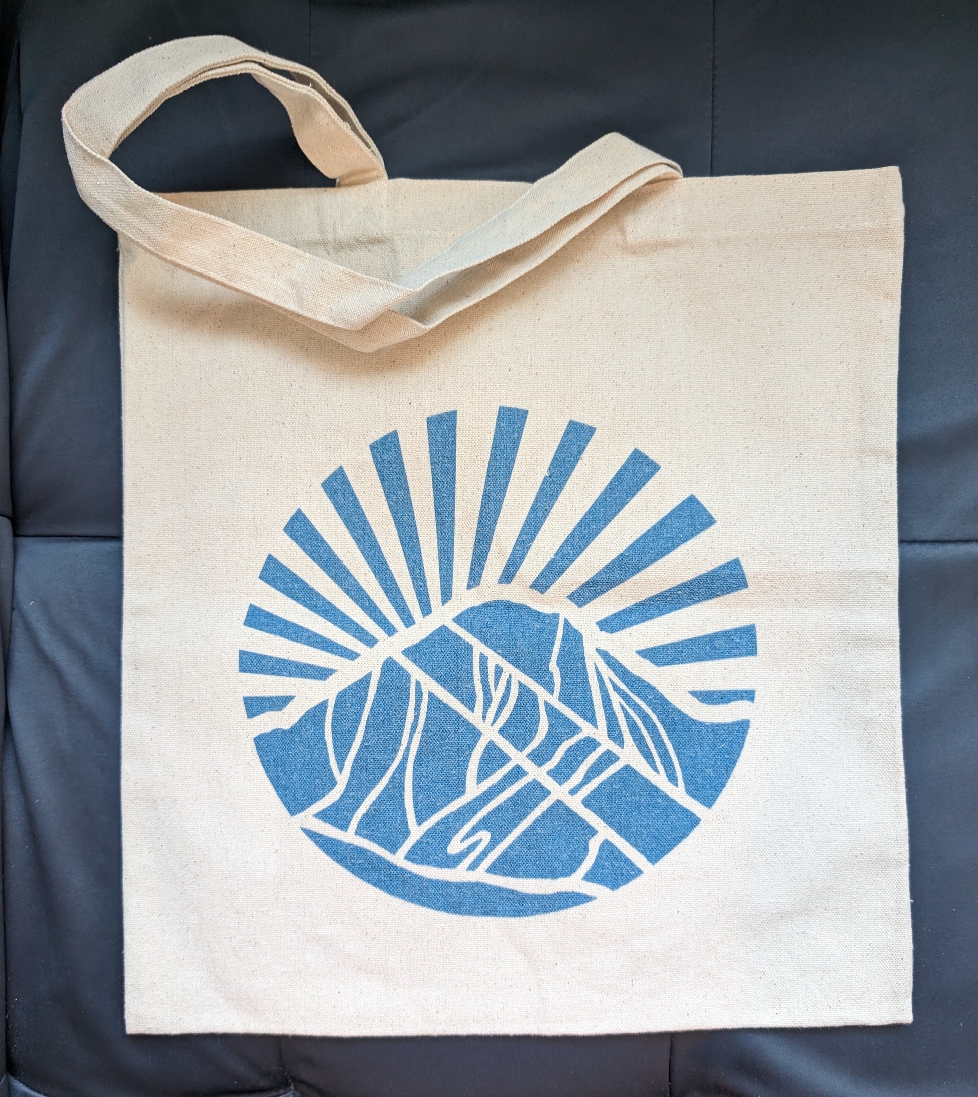 Canvas tote bag with blue icon of a mountain with light rays radiating from the mountain to the sky